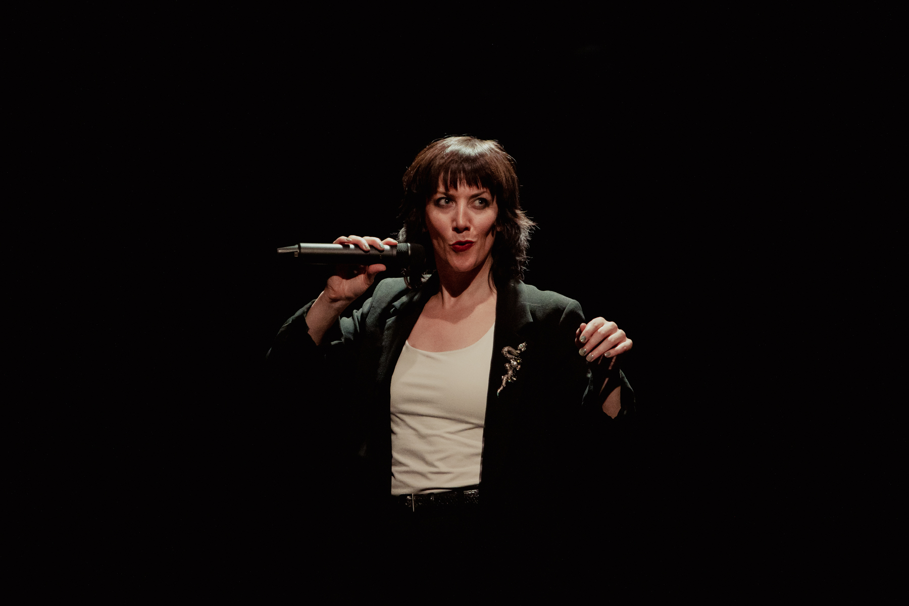 Actress Deborah Pugh is wearing a white vest and a black blazer with a snake brooch. She is holding the mic next to her lips with her right hand. Photo taken by Camilla Adams.