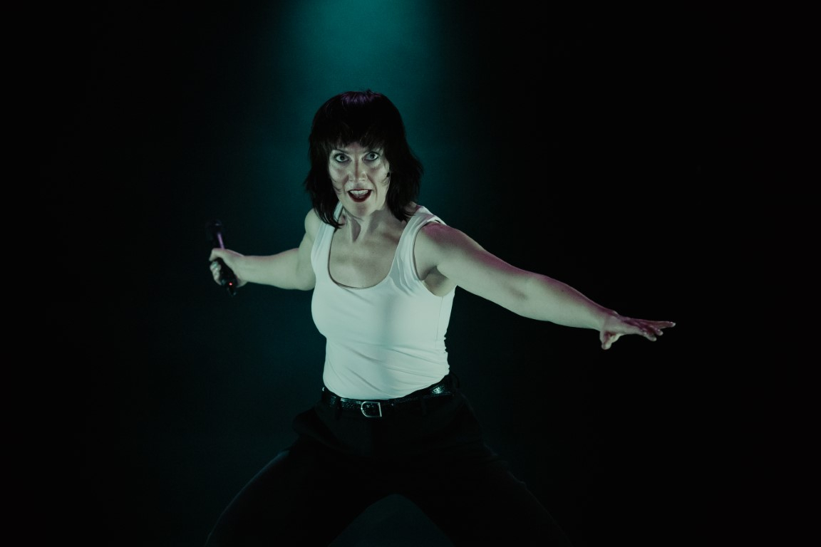 Actress Deborah Pugh is wearing a white vest and black trousers. She is facing directly at the camera and smiling. She has the mic in her right hand behind her body, and her left hand is towards the front. There is a pale green light on her.