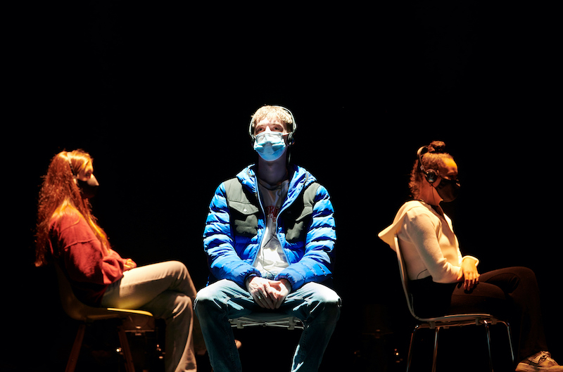 Under The Mask production photographs. People on a stage, spaced apart wearing headphones and a mask.
