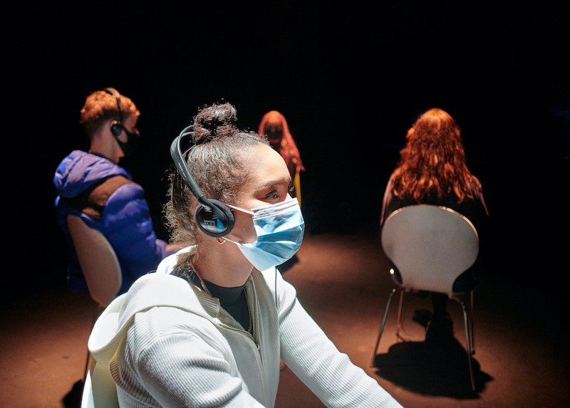 Under The Mask production photographs. People on a stage, spaced apart wearing headphones and a mask.