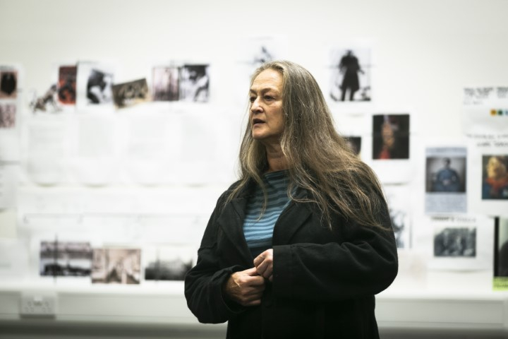 Liz looks to the side with both hands clasped in front of her. In the background is blurred set and costume images on the rehearsal wall.