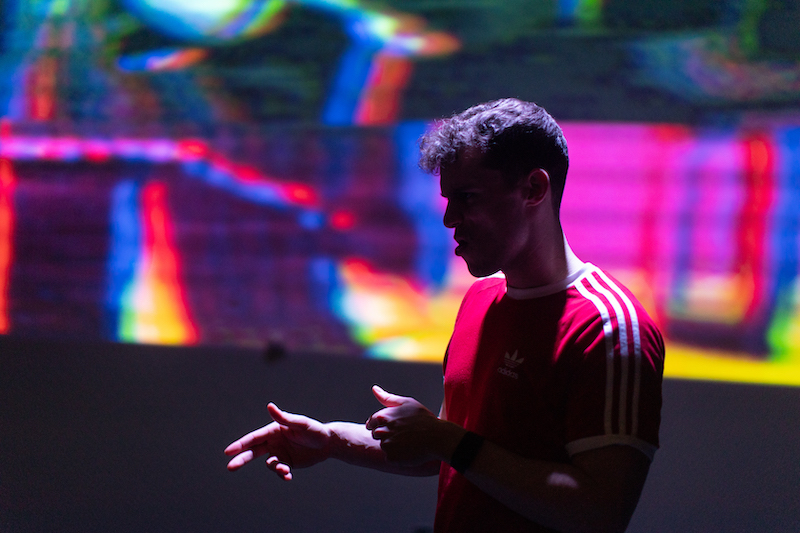 Y'MAM Production pictures. Sam Churchill in red addidas top in front of a projection with purple & yellow tones.
